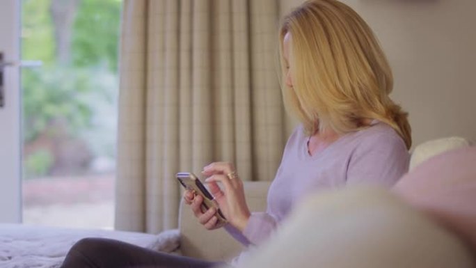 在家中的成熟女人坐在沙发上使用手机发短信或社交媒体