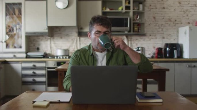 白人男子坐在餐桌旁，用笔记本电脑在家喝咖啡