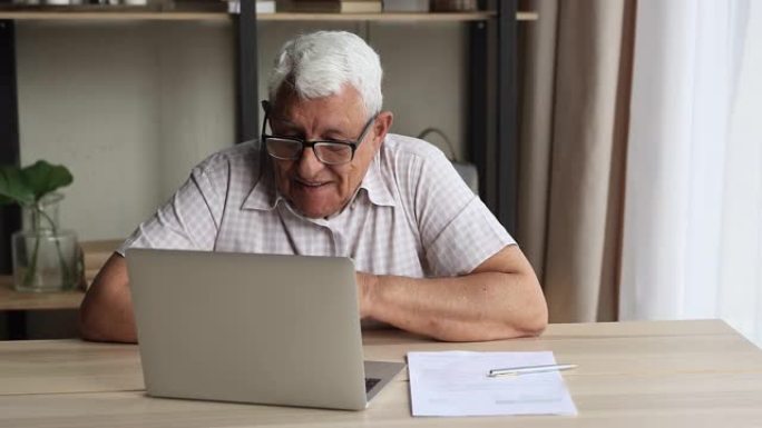 高年级男子坐在桌旁使用笔记本电脑进行视频通话
