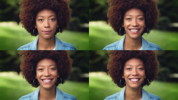 漂亮的卷发女性享受新鲜的空气和自然，与世界分享她灿烂的笑容。一位年轻的黑人妇女在绿色公园里看着相机大