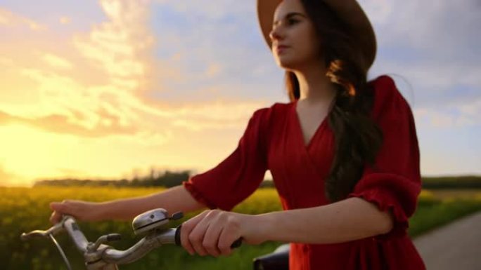 追踪，一个穿着红色连衣裙的黑发女人的特写镜头，她骑着自行车，周围是黄色的金鱼草，背景是日落