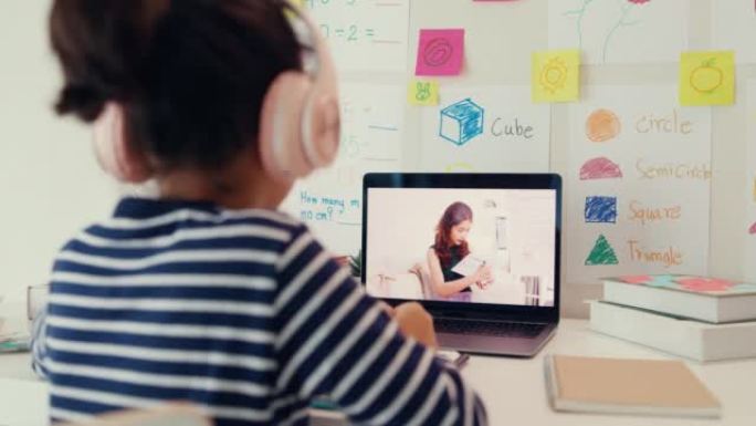 亚洲蹒跚学步的女孩戴着耳机坐在带记事本的书桌前使用魔术笔专注于写作做作业听老师在家从笔记本电脑屏幕上