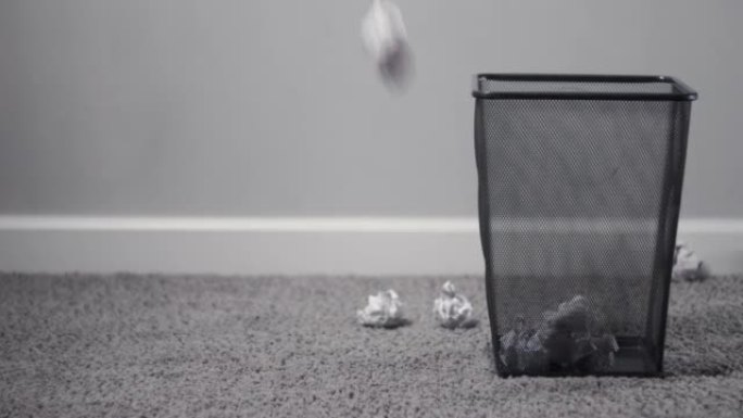 白色皱巴巴的纸球从无处掉落在灰色地毯地板上的黑色金属废纸篮中及其周围