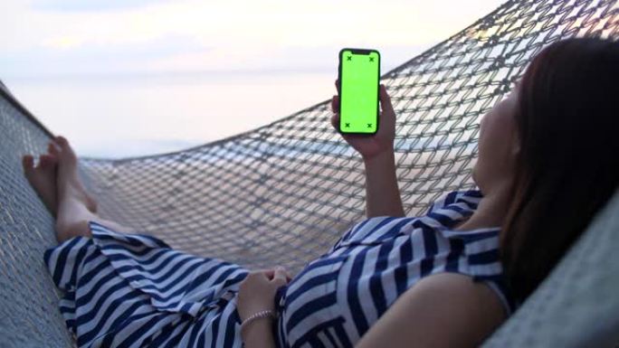 年轻女孩躺在海滩上的吊床上时使用智能手机