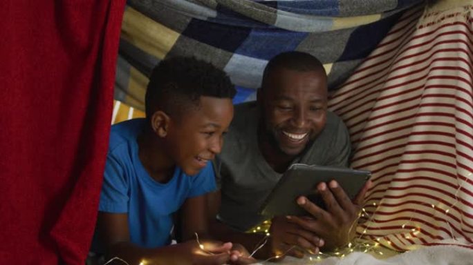 快乐的非裔美国人父子使用平板电脑，躺在毯子营地大笑