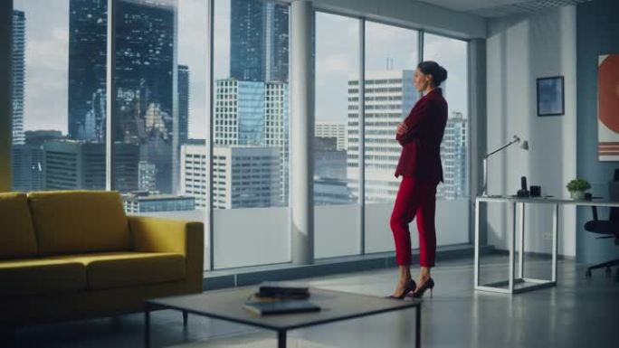 成功的体贴的高加索女商人穿着完美的红色西装站在办公室，看着窗外的大城市。自信的女性公司首席执行官管理