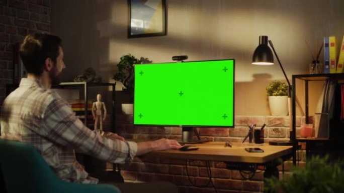 办公室员工在家工作，在带有绿屏模拟显示器的台式计算机上进行视频通话。男人坐在客厅里，晚上与朋友和家人