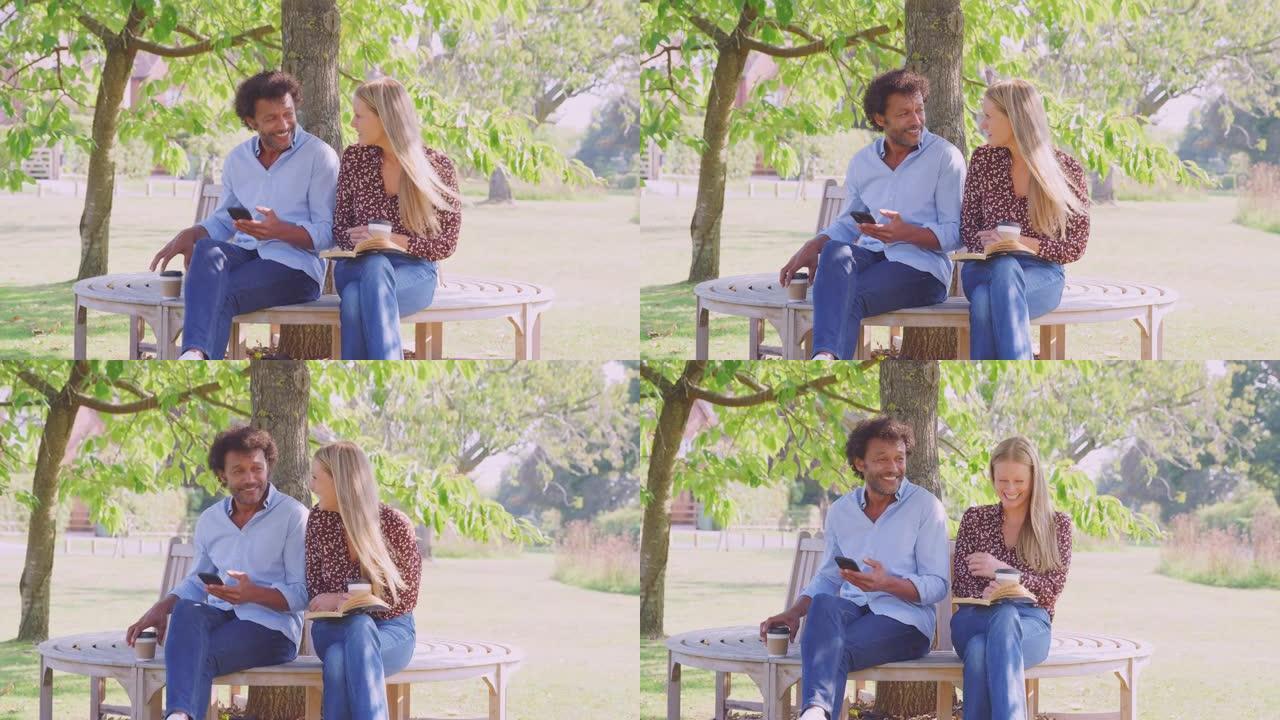 成熟的夫妇放松坐在公园长椅上树下看书和检查手机