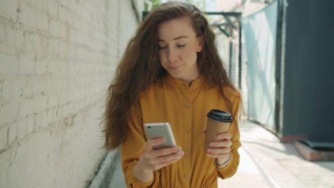 美丽的女孩走在城市街道上喝咖啡和使用智能手机的肖像