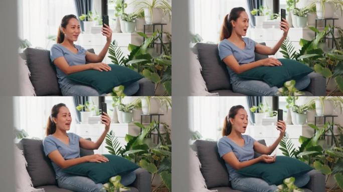年轻的亚洲女性在沙发上休闲穿着使用智能手机举起手视频通话打个招呼，并在家里的客厅室内植物中与家人和朋