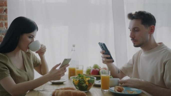 夫妻在家里的厨房里吃早餐，默默地使用智能手机