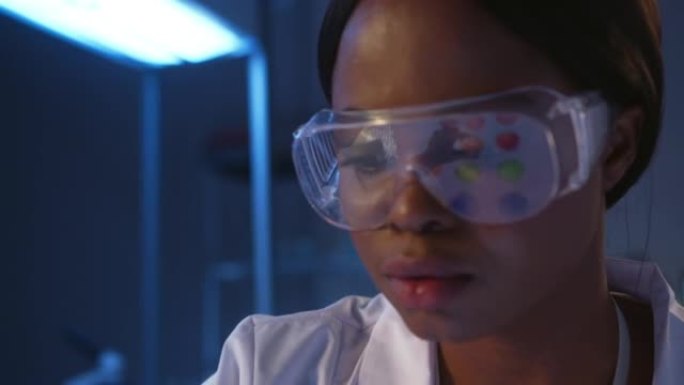 一位戴着防护眼镜的严肃的深色皮肤微生物学家在实验室工作时的特写镜头，我们在眼镜的桌子上看到小培养皿的