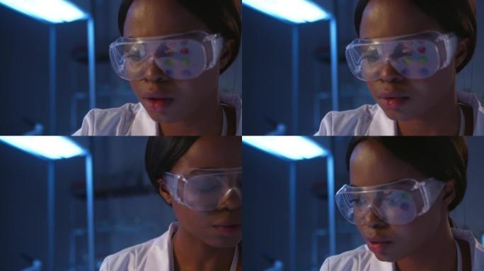 一位戴着防护眼镜的严肃的深色皮肤微生物学家在实验室工作时的特写镜头，我们在眼镜的桌子上看到小培养皿的