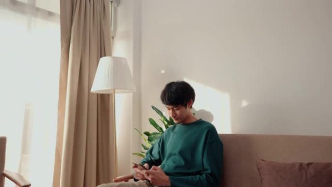 亚洲帅哥坐在沙发上在线智能手机聊天。
