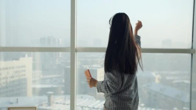 亚洲女人戴着耳机跳舞的慢动作在全景窗户背景下在家玩耍