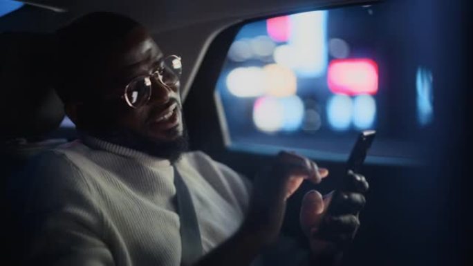 戴着眼镜的快乐黑人晚上坐出租车后座回家。英俊的男性使用智能手机，在城市街道上开着霓虹灯的汽车时微笑。