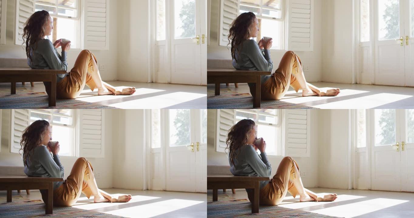 体贴的白人妇女坐在阳光明媚的小屋的地板上喝咖啡，看着窗外