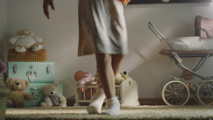 穿着睡裙和舒适袜子的小女孩的电影特写镜头正试图用芭蕾舞娃娃在托儿所的地毯上尝试古典芭蕾舞的步骤。舞蹈