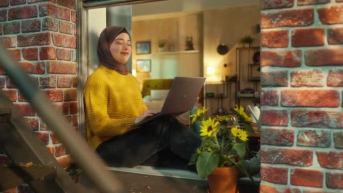 一位戴着头巾的年轻穆斯林女性坐在窗台上在笔记本电脑上工作的肖像。商务办公室员工在家远程工作。从窗外看
