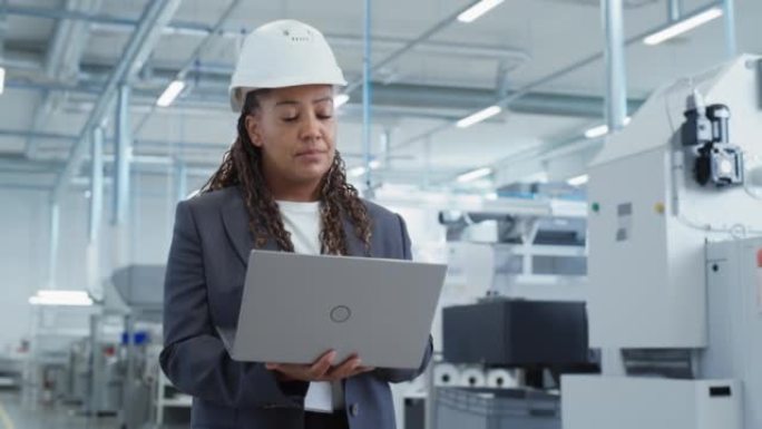 一位戴着安全帽的黑人女工程师在电子制造工厂站着并使用笔记本电脑的肖像。处理日常任务和研发数据的技术人