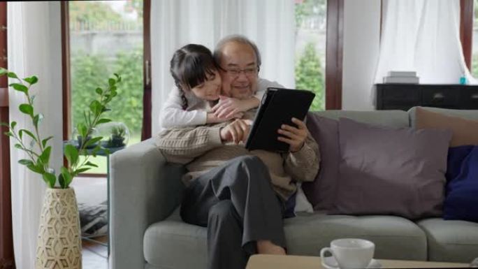 亚洲老前辈祖父母的坦率与孩子们在家庭中的亲密关系中与家庭中的平板电脑上的技术一起玩耍和观看。年轻女孩