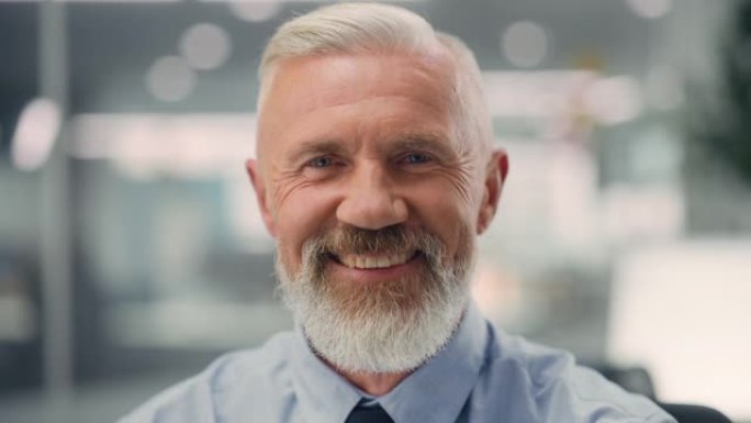 一位自信快乐的高级男性穿着蓝色衬衫，看着相机，真诚迷人地微笑着。成功经验丰富的老人在不同的公司办公室