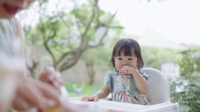 亚洲母亲喂养她的女婴坐在公园的高脚椅上。