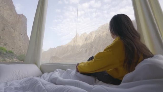 年轻女子的后视图，穿着黄色的衣服，包裹在温暖的毯子，而在早晨醒来，望向窗外的湖。种族多样化的旅行者概