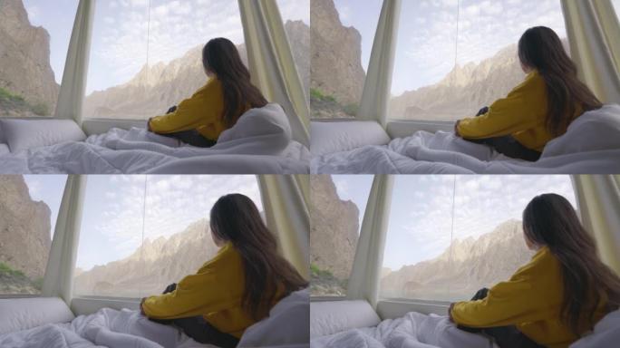年轻女子的后视图，穿着黄色的衣服，包裹在温暖的毯子，而在早晨醒来，望向窗外的湖。种族多样化的旅行者概
