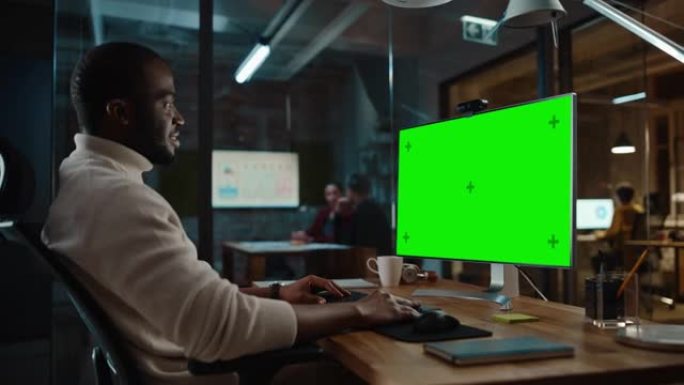 英俊的黑人非裔美国人项目经理正在繁忙的创意办公室中使用带有绿屏模型显示的台式计算机进行视频通话。男性