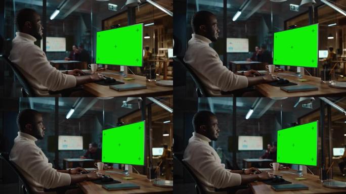 英俊的黑人非裔美国人项目经理正在繁忙的创意办公室中使用带有绿屏模型显示的台式计算机进行视频通话。男性