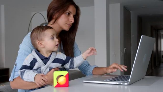 年轻的母亲在家里的一次商务会议上使用笔记本电脑，同时将婴儿抱在膝盖上