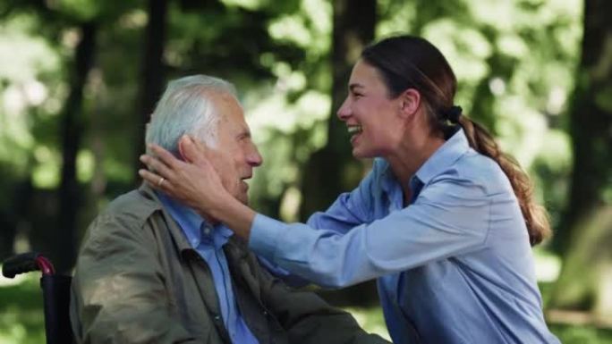 电影拍摄的快乐孙女在轮椅上给祖父以情感的吻，以此作为绿色公园中爱与尊重的标志。生活观念，祖父母，世代