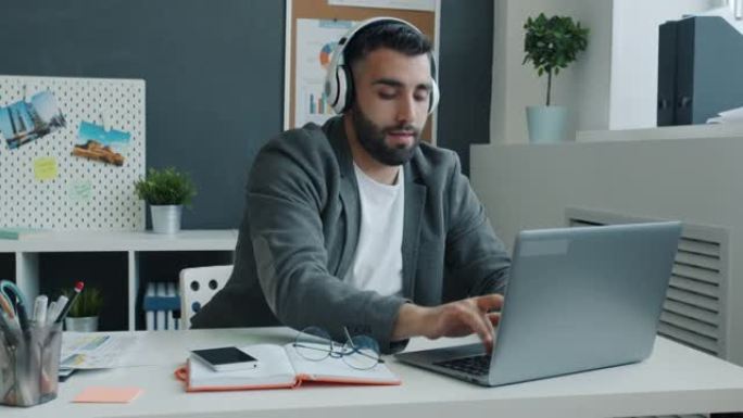 有趣的商人戴着耳机跳舞，用笔记本电脑坐在办公室的桌子上工作