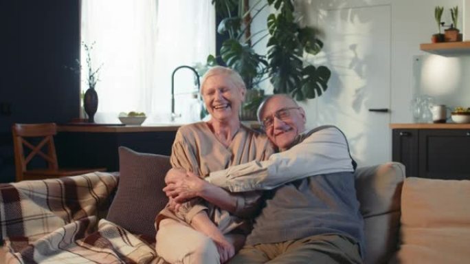 快乐开朗的退休浪漫60年代高级丈夫拥抱妻子微笑，坐在舒适客厅的沙发上聊天。