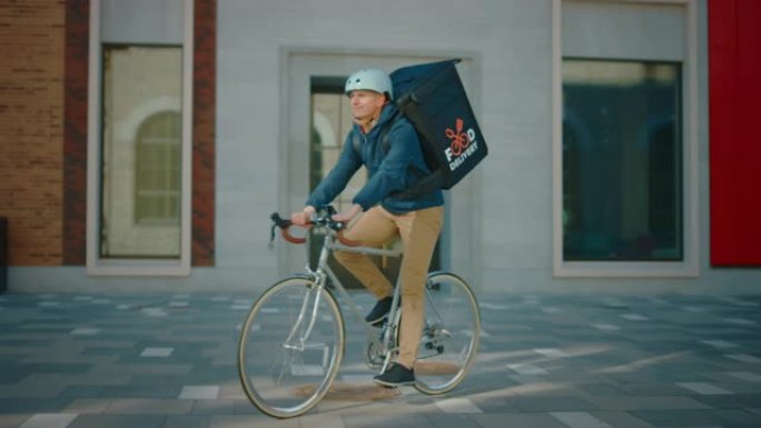 穿着保暖背包的英俊快乐送餐男子骑着自行车穿过时尚的现代市区。微笑的快递员安全交付餐厅订单的肖像。慢动