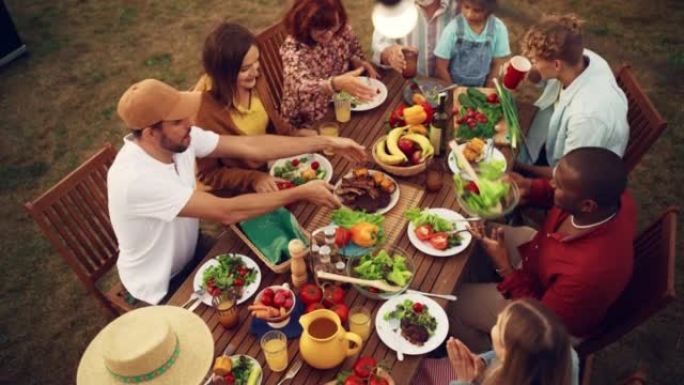 家人和朋友在家里庆祝的俯视图。各种各样的儿童，成人和老年人坐在桌子旁，进行有趣的交谈。吃烧烤和蔬菜。