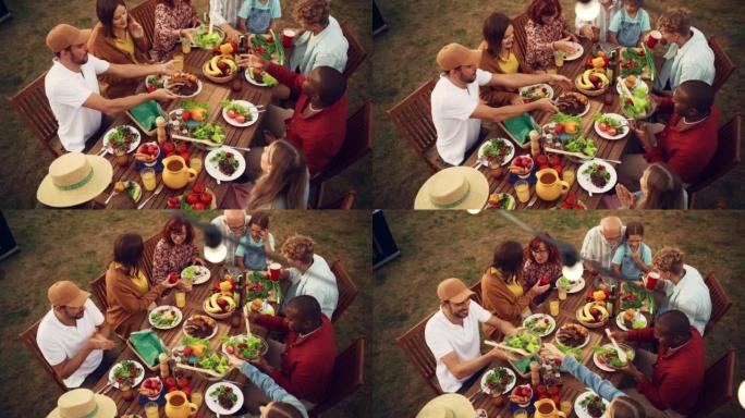家人和朋友在家里庆祝的俯视图。各种各样的儿童，成人和老年人坐在桌子旁，进行有趣的交谈。吃烧烤和蔬菜。