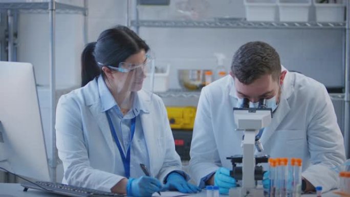 男女实验室工作人员使用显微镜进行研究，并在计算机上记录结果