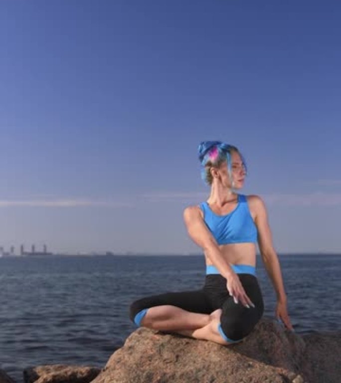 一位迷人的健美瑜伽老师，穿着蓝黑色的瑜伽服，一头蓝色的头发，盘腿坐在一块岩石上，在白天的天空和大海的