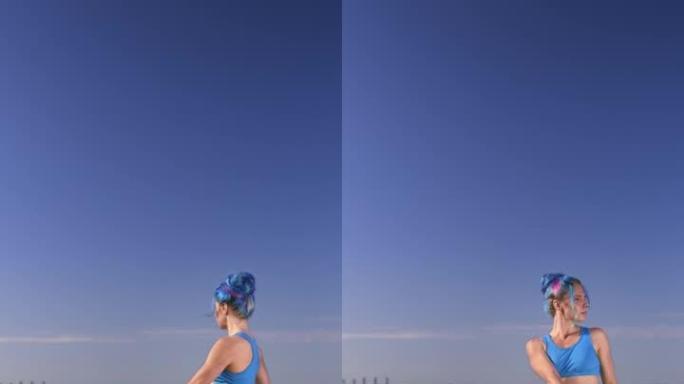 一位迷人的健美瑜伽老师，穿着蓝黑色的瑜伽服，一头蓝色的头发，盘腿坐在一块岩石上，在白天的天空和大海的