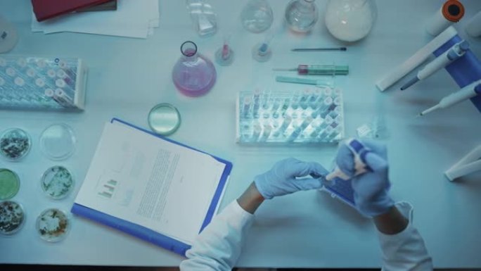 在现代实验室的桌子后面，戴着蓝色橡胶手套的医学研究科学家的头部摄像机镜头。医生使用试管，比较样本并写