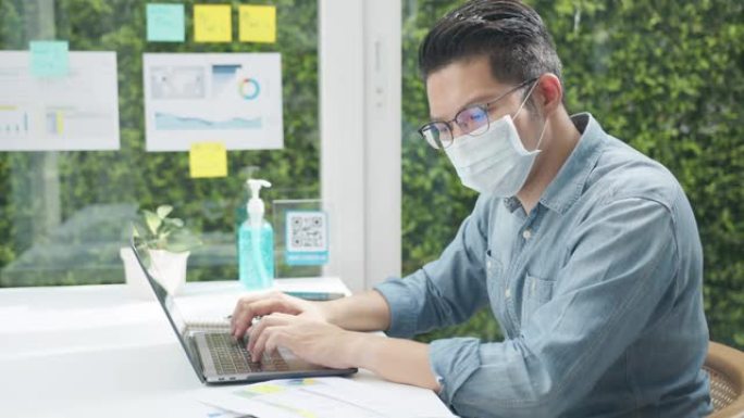 亚洲官员男子戴着口罩在办公室用笔记本电脑工作。冠状病毒危机后的新常态、社交距离措施和工作生活方式。