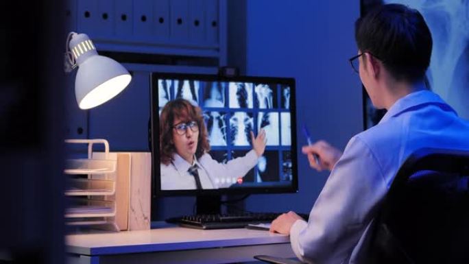 亚洲男性医生视频电话给白人男性教育医生关于视频会议上骨骼的研究x射线，而在深夜工作的家中通过计算机进