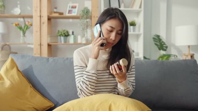 生病的年轻亚洲妇女拿着药坐在沙发上使用智能手机通话在家咨询医生。