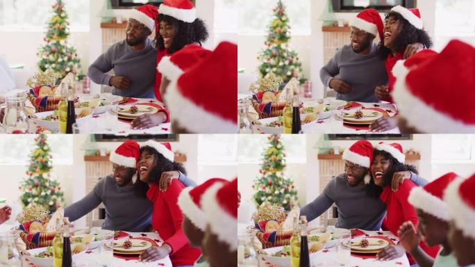 戴着圣诞老人帽子的非洲裔美国夫妇坐在餐桌上吃lunc时微笑和敬酒