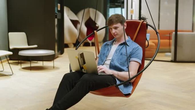 年轻人使用笔记本电脑坐在创意办公室休息室的秋千椅上