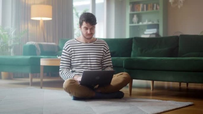 英俊的成年男子使用笔记本电脑，同时坐在舒适时尚的公寓的客厅地板上。有魅力的男人正在网上购物，在流媒体