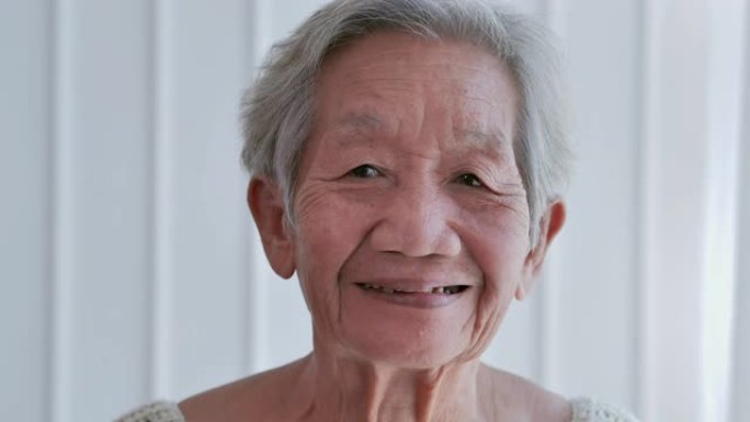 资深的白发老妇在家里看着相机。心情愉快，心情愉快，笑容灿烂的老年妇女。快乐的老奶奶，在家里或养老院露