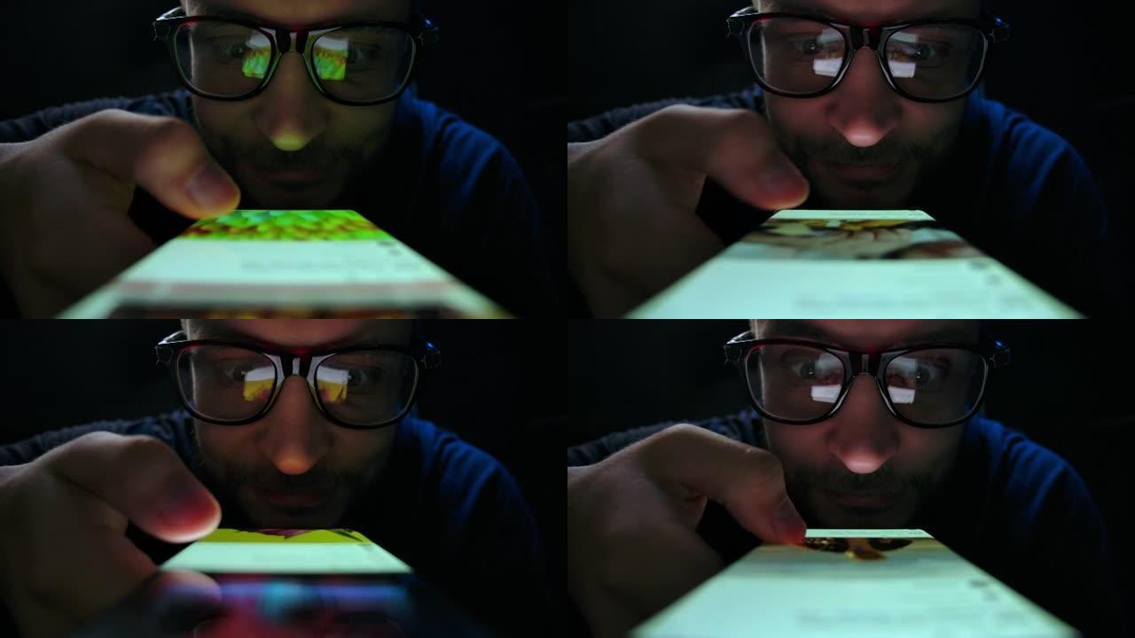 戴眼镜的快乐男人看手机，浏览社交网络特写。有趣而轻松的年轻成年男性晚上在手机上阅读应用程序。手机触摸